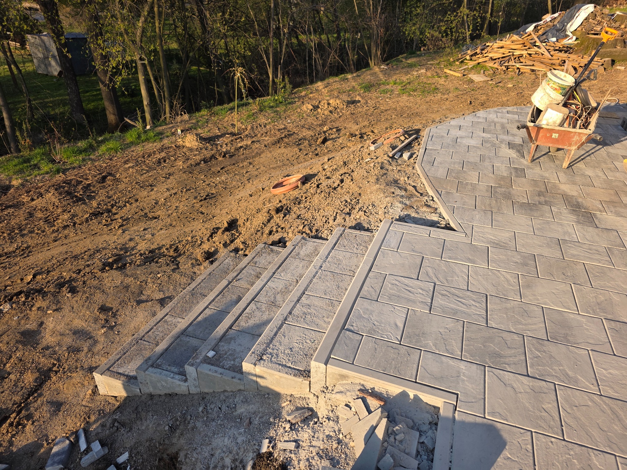 Ułożone schody z kostki brukowej na zboczu, w tle ziemia i drzewa, na górze wózek z narzędziami. Widok z góry, w słoneczny dzień.