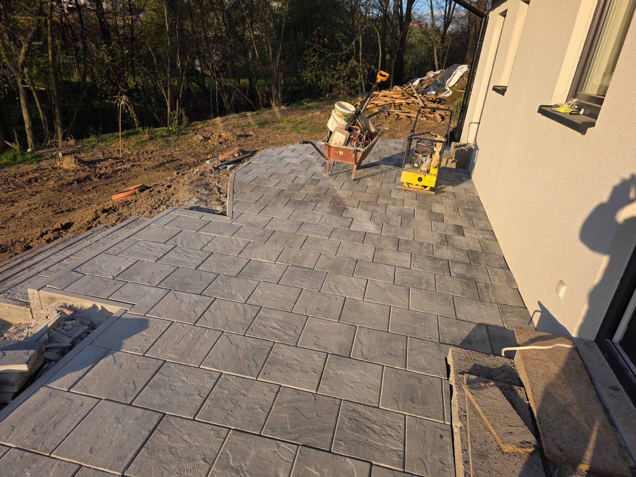 Ułożona szara kostka brukowa na tarasie domu, z widocznymi narzędziami (taczka, ubijarka) i materiałami budowlanymi w tle, w słoneczny dzień.