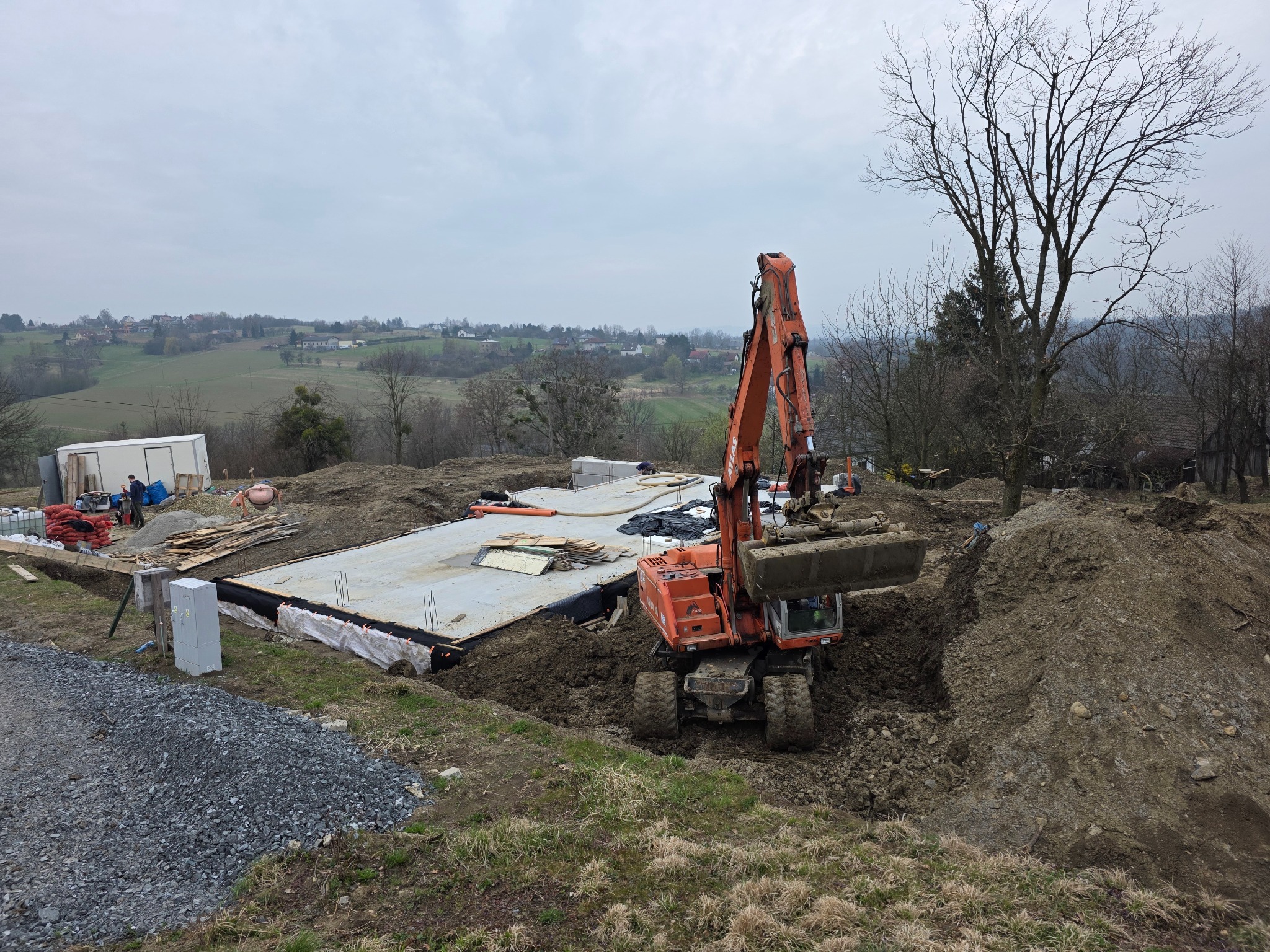 Pomarańczowa koparka przy wykopie pod fundamenty na tle krajobrazu. Widoczny zarys fundamentu i przygotowane zbrojenie. Prace ziemne w toku.