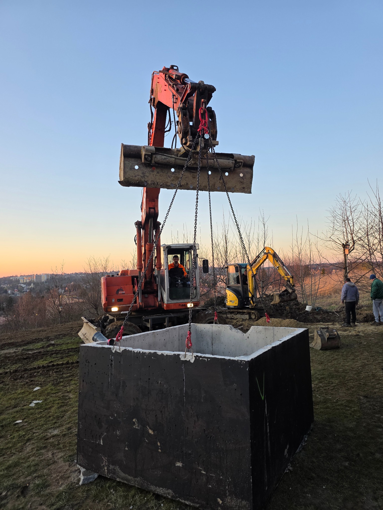 Czerwona koparka z łyżką zawieszoną na łańcuchach, unosi betonowy element. W tle druga koparka i osoby. Widok na miasto o zachodzie słońca.