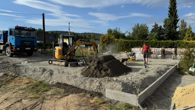 Przygotowanie terenu pod kostkę: mini koparka, ubijarka, pryzma ziemi i palety z kostką na żwirowym podłożu. Widoczny fragment ułożonego obrzeża.