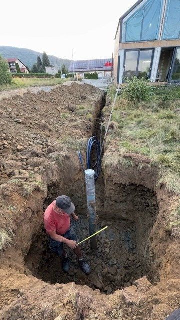 Głęboki wykop w ziemi z pionowo umieszczoną rurą i niebieskimi kablami; mężczyzna mierzy głębokość wykopu żółtą miarką, w tle dom z panelami słonecznymi.