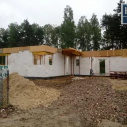 Wznoszenie ścian parterowego domu z bloczków, widoczne drewniane elementy szalunku i podparcia, palety bloczków za ogrodzeniem oraz logo firmy w prawym górnym rogu.