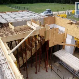 Budowa domu murowanego: widok z g&oacute;ry na strop Teriva zbrojony stalą, szalunki schod&oacute;w z desek i płyt OSB, pracownik w niebieskim kombinezonie układa elementy stropu, w tle samoch&oacute;d z bagażnikiem...