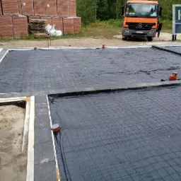 Przygotowany fundament pod budowę domu, z widoczną izolacją, siatką zbrojeniową, elementami kanalizacji, ułożonymi cegłami i zaparkowaną ciężarówką w tle.