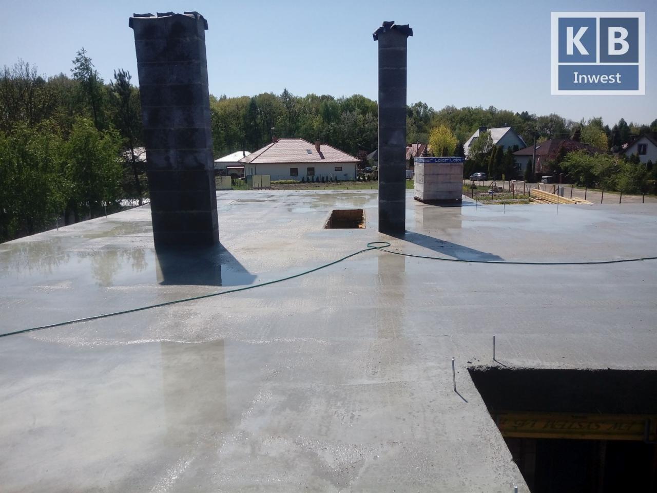 Wylany, mokry strop betonowy z dwoma kominami w stanie surowym, widok z góry na tle zabudowy jednorodzinnej i drzew. Na stropie widoczny zielony kabel.