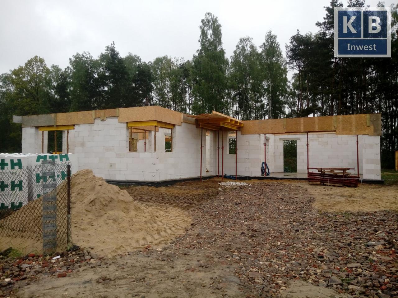 Wznoszenie ścian parterowego domu z bloczków, widoczne drewniane elementy szalunku i podparcia, palety bloczków za ogrodzeniem oraz logo firmy w prawym górnym rogu.