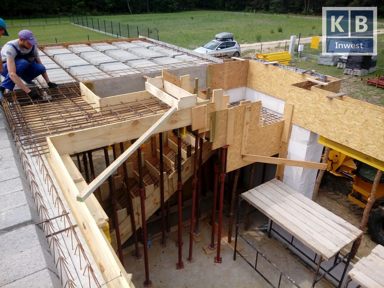 Budowa domu murowanego: widok z góry na strop Teriva zbrojony stalą, szalunki schodów z desek i płyt OSB, pracownik w niebieskim kombinezonie układa elementy stropu, w tle samochód z bagażnikiem...