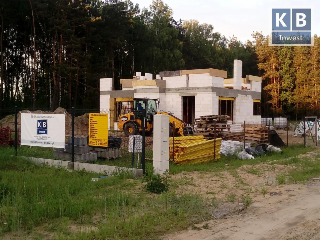 Wznoszenie domu z białych bloczków, widoczny żółty traktor i tablica informacyjna na ogrodzeniu budowy, las w tle.