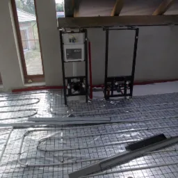 Stelaże podtynkowe marki Koło zamontowane na podłodze z rozłożonym ogrzewaniem podłogowym w trakcie prac instalacyjnych, widoczne rury grzewcze w siatce, surowy stan wykończenia wnętrza.