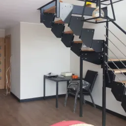 Nowoczesne schody metalowe z drewnianymi stopniami w jasnym wnętrzu, widok z dołu na konstrukcję i część pokoju z meblami.