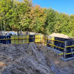 Szczegółowe ujęcie szalunków fundamentowych z żółtymi panelami i metalowymi rozpórkami, na tle zielonego lasu i błękitnego nieba, częściowo zasypane ziemią.