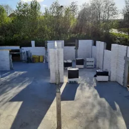 Wczesny etap budowy domu z betonu komórkowego, widoczne ściany nośne i działowe, palety z bloczkami H+H na betonowej płycie fundamentowej, zieleń drzew w tle.