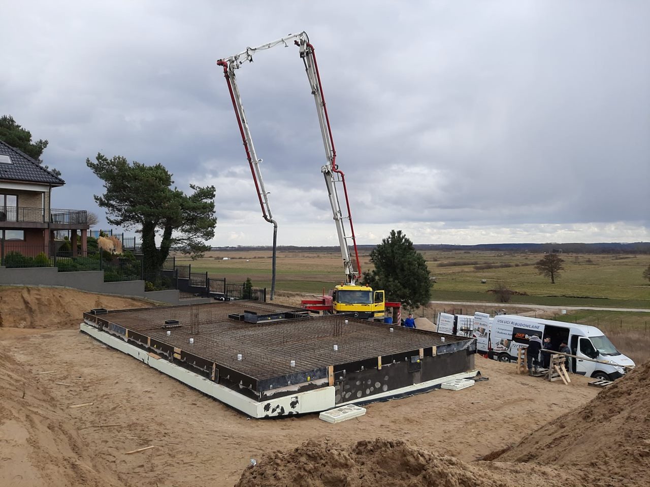 Wylewanie betonu na zbrojony fundament domu jednorodzinnego za pomocą pompy do betonu, widoczny budynek w tle i samochód dostawczy z logo firmy budowlanej.