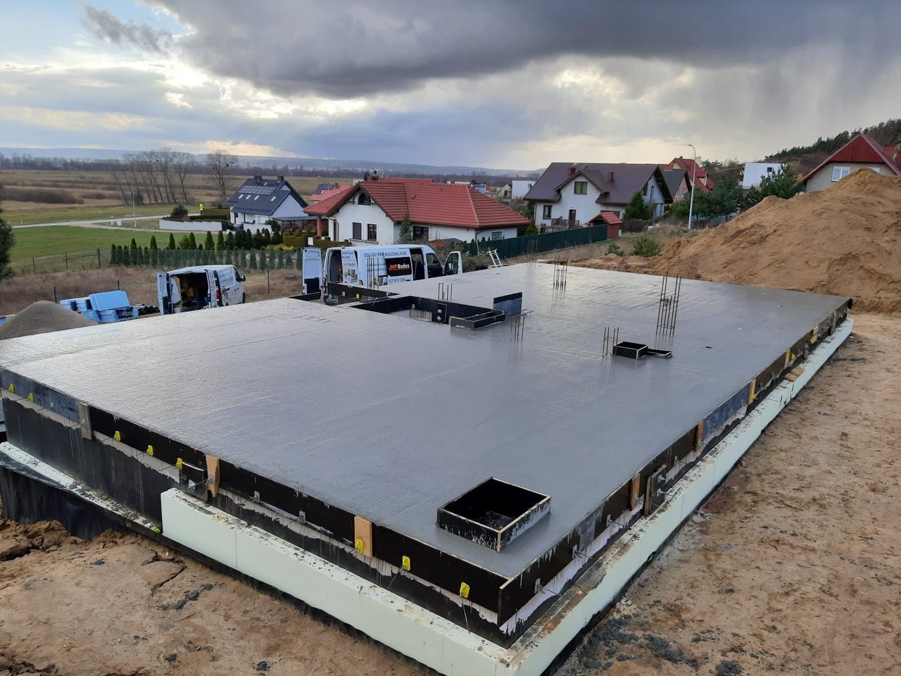 Świeżo wylana płyta fundamentowa z widocznymi otworami na instalacje, zabezpieczona izolacją termiczną, na tle zabudowy jednorodzinnej i pochmurnego nieba.