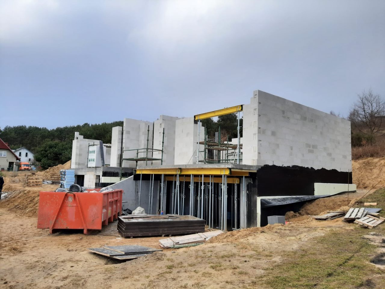 Surowy stan domu z betonu komórkowego w trakcie budowy, widoczne szalunki i rusztowania, teren budowy z kontenerem i materiałami.