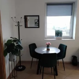 Jasne wnętrze pokoju z okrągłym stołem, czterema ciemnozielonymi krzesłami, wazonem z różowymi kwiatami na koronkowej serwetce, w tle okno z szarą roletą, na parapecie doniczka z trawą, obok...