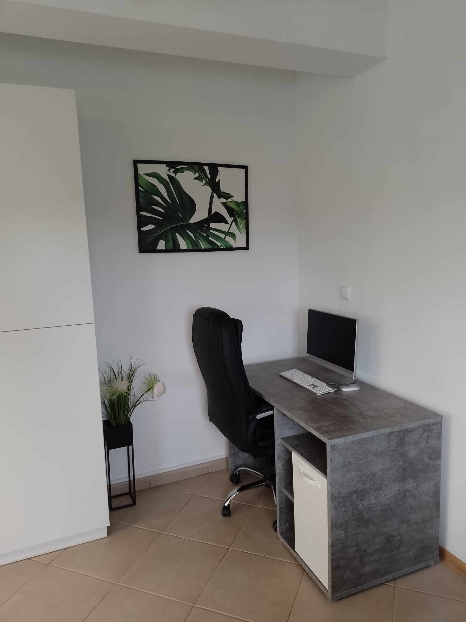 Biuro domowe z szarym biurkiem, komputerem, czarnym krzesłem obrotowym i obrazem z motywem roślinnym na białej ścianie, roślina w czarnej doniczce obok białej szafki.