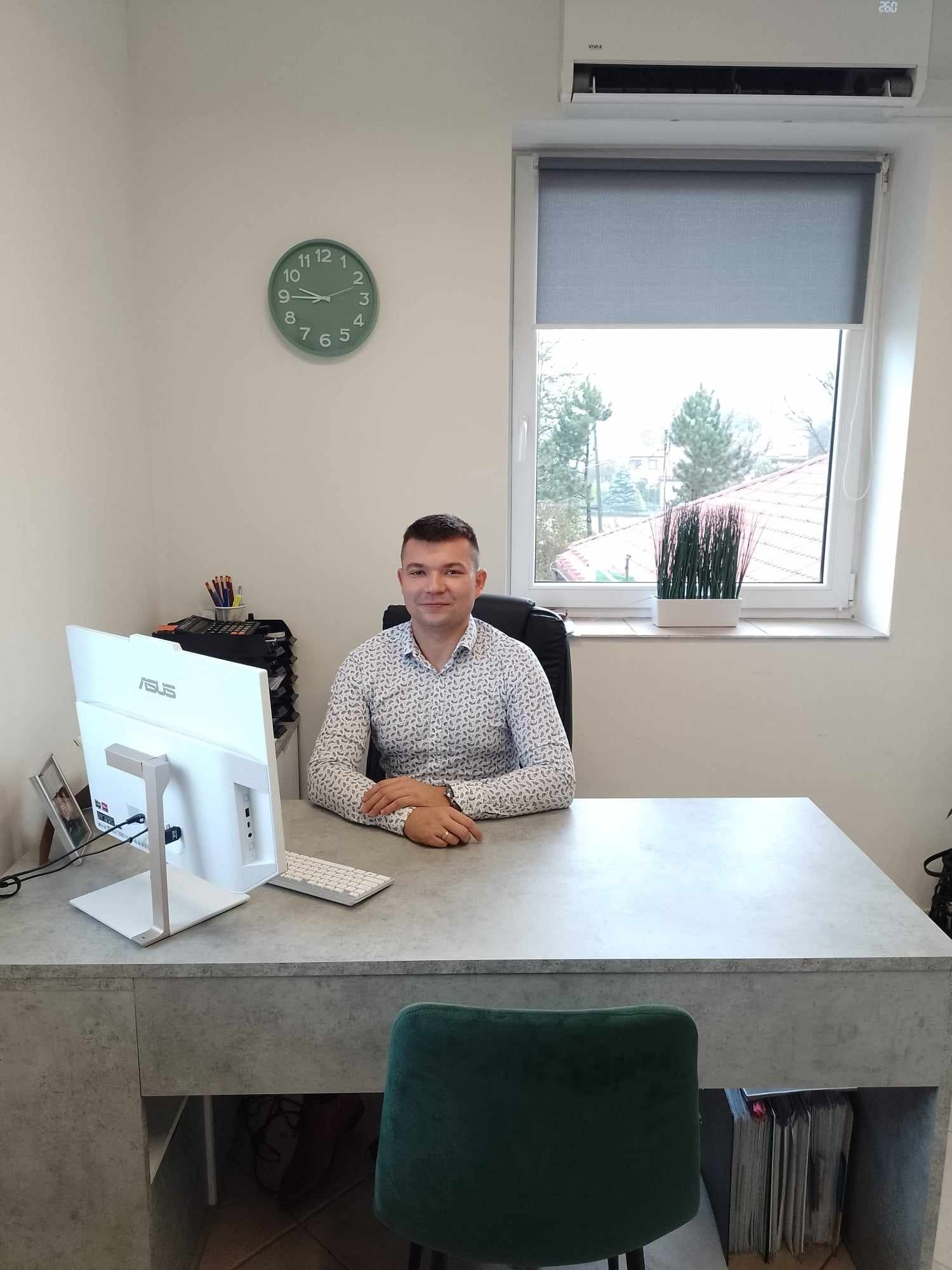 Młody doradca finansowy siedzi za biurkiem w jasnym biurze, z zielonym zegarem na ścianie i klimatyzatorem nad oknem.