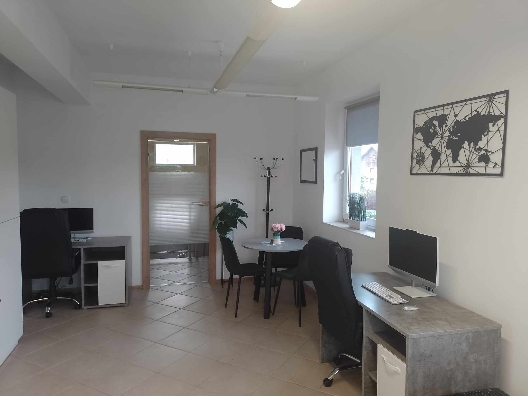 Jasne biuro z dwoma stanowiskami pracy z komputerami, stołem konferencyjnym z kwiatami i dekoracją ścienną w postaci mapy świata, widok na sąsiednie pomieszczenie przez otwarte drzwi.