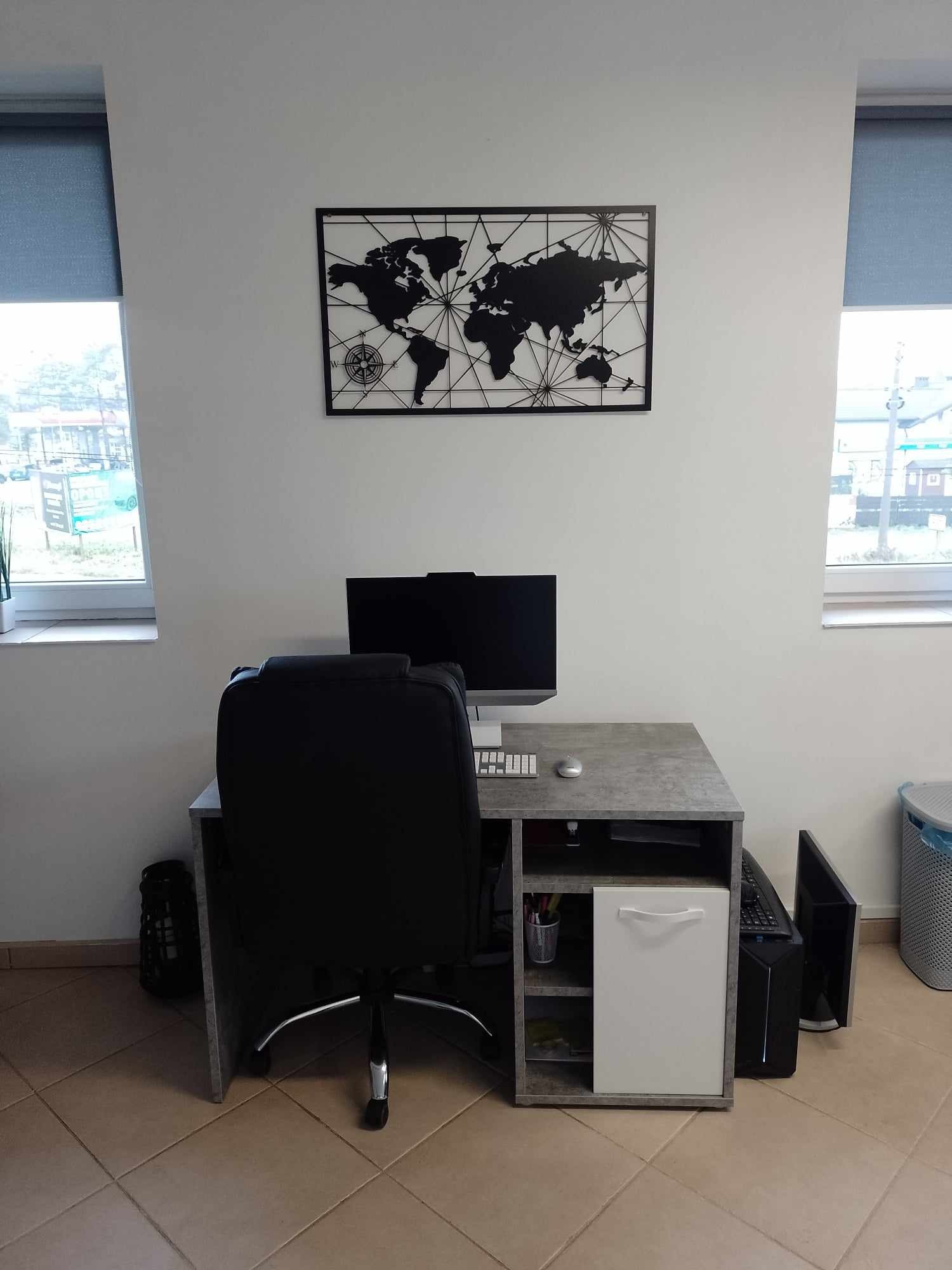 Biuro domowe z szarym biurkiem, czarnym krzesłem obrotowym i mapą świata na ścianie, widok z przodu, na biurku komputer stacjonarny z monitorem i klawiaturą.
