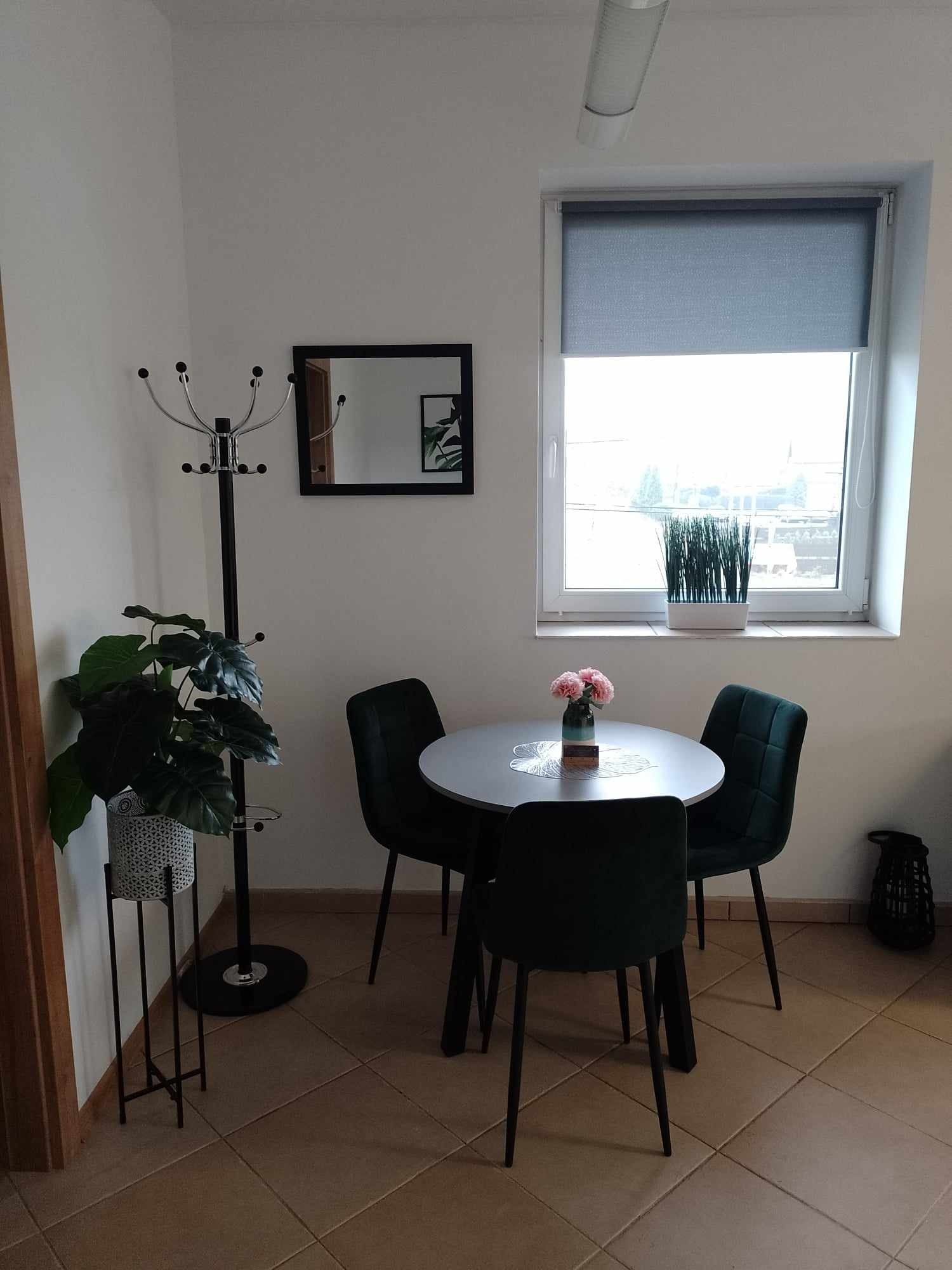 Jasne wnętrze pokoju z okrągłym stołem, czterema ciemnozielonymi krzesłami, wazonem z różowymi kwiatami na koronkowej serwetce, w tle okno z szarą roletą, na parapecie doniczka z trawą, obok...