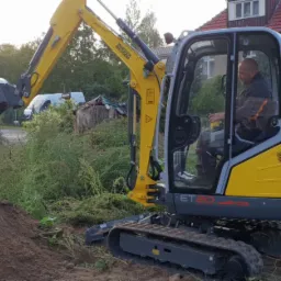 Żółto-szara mini koparka Wacker Neuson ET20 z operatorem w kabinie podczas pracy w terenie z ziemią i zielenią w tle.