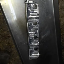 Metalowa listwa z otworami, prawdopodobnie część silnika samochodowego, leżąca na metalowej tacy warsztatowej, widoczne ślady użytkowania i odbicia światła.