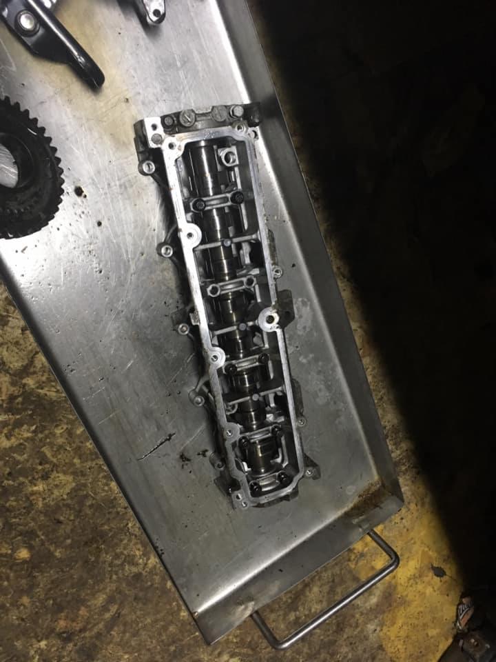 Zdemontowana głowica silnika z wałkami rozrządu na metalowej tacy warsztatowej, widoczne ślady oleju i zabrudzenia.