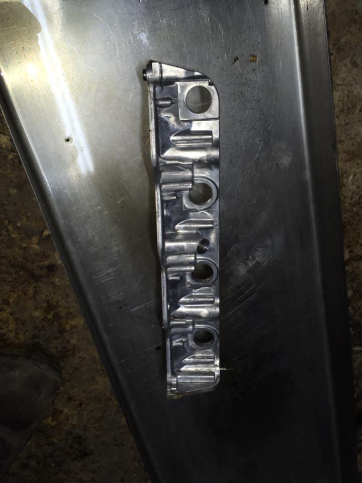 Metalowa listwa z otworami, prawdopodobnie część silnika samochodowego, leżąca na metalowej tacy warsztatowej, widoczne ślady użytkowania i odbicia światła.