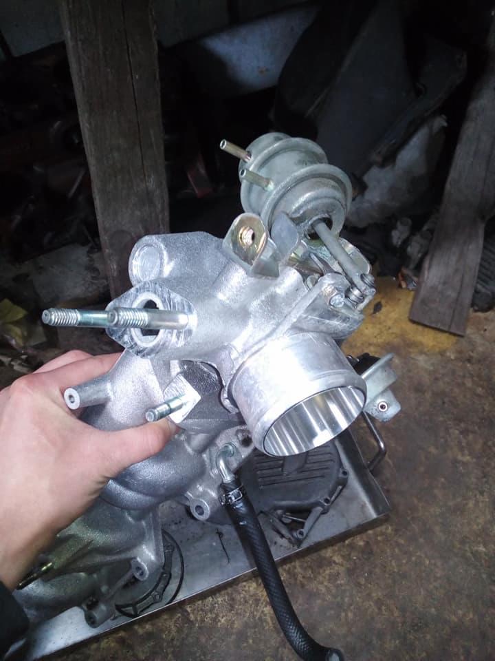 Oczyszczony, srebrny kolektor dolotowy z przepustnicą i regulatorem ciśnienia paliwa, trzymany w dłoni, widoczne śruby i przewód paliwowy, na tle warsztatu.