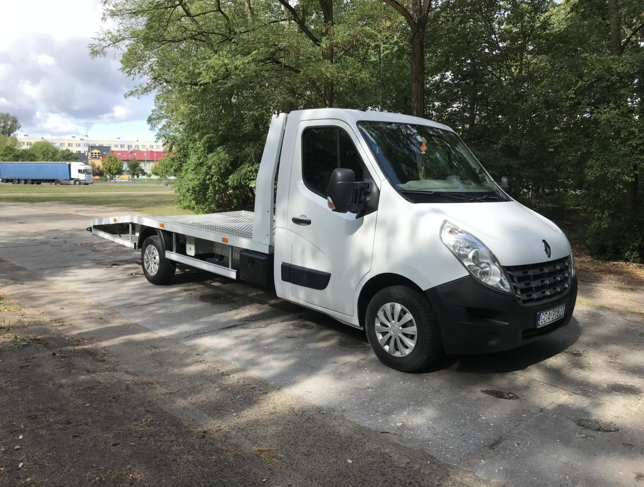 Biała laweta Renault Master na betonowym placu, z widoczną tablicą rejestracyjną i drzewami w tle.