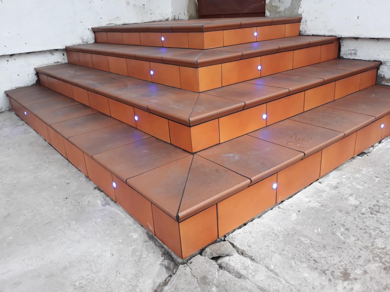 Zewnętrzne schody wejściowe wyłożone terakotą z wbudowanym oświetleniem LED, widoczne pęknięcia w betonie u podstawy.