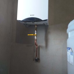 MK-INSTAL Michał Kaczmarski - Biały wiszący kocioł gazowy marki Termet z podłączeniem miedzianymi rurami i żółtym zaworem, zamontowany na surowej ścianie.