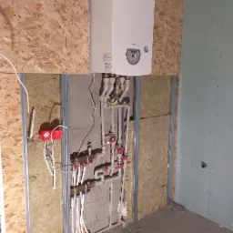 Biały wiszący piec gazowy Termet z podłączonymi rurami instalacji grzewczej i wodnej w surowym pomieszczeniu z płyt OSB i gipsowo-kartonowych, widoczne zawory i kształtki.