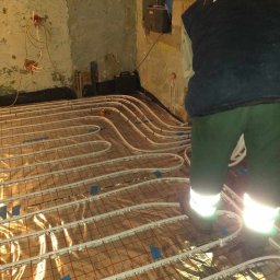 MK-INSTAL Michał Kaczmarski - Instalacja ogrzewania podłogowego: białe rurki ułożone na folii z czerwoną siatką, widoczne nogi instalatora w zielonych spodniach z odblaskami, surowe ściany w tle.