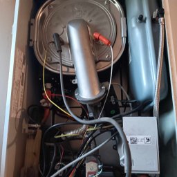 MK-INSTAL Michał Kaczmarski - Wnętrze kotła gazowego z widocznymi elementami instalacji, w tym srebrną obudową palnika i zbiornikiem wyrównawczym, liczne przewody i kable wewnątrz urządzenia.