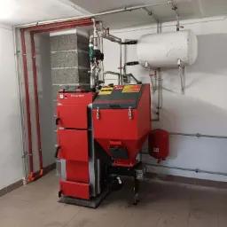 Czerwono-szary piec na paliwo stałe marki DEFRO z zbiornikiem buforowym i instalacją hydrauliczną w kotłowni z bloków betonowych i ceramicznej podłodze.