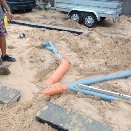 MK-INSTAL Michał Kaczmarski - Instalacja rur kanalizacyjnych w piasku, widoczne kształtki z tworzywa sztucznego, poziomica, obrzeża chodnikowe, przyczepa i samochód w tle.