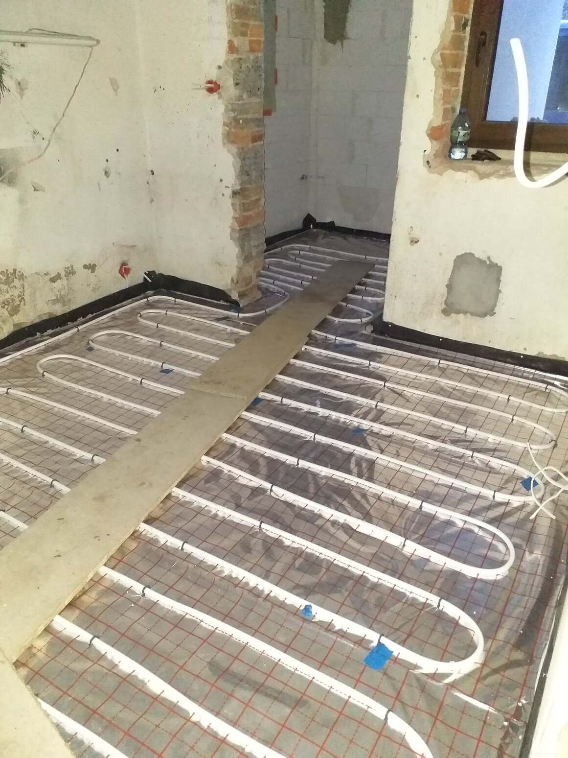 Montaż ogrzewania podłogowego w surowym wnętrzu, rury grzewcze ułożone na folii z czerwoną siatką, zabezpieczone deską, widoczne fragmenty ścian w stanie deweloperskim i okno.