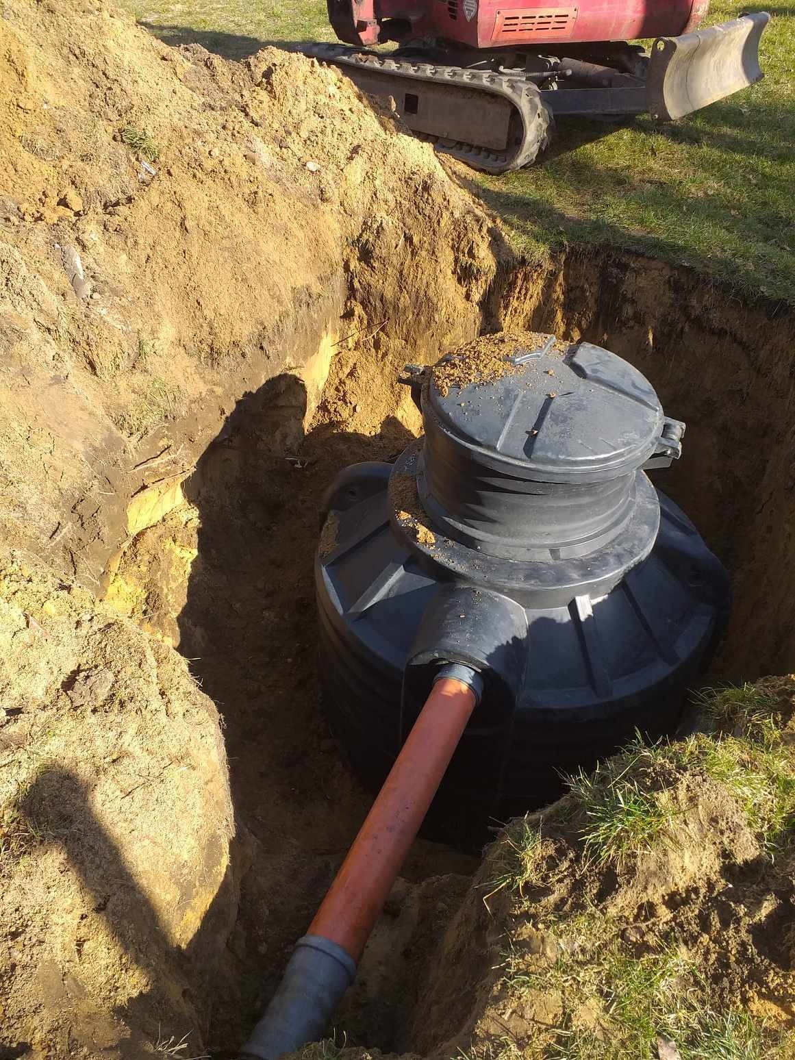 Czarny zbiornik bezodpływowy z rurą w wykopie ziemnym, z widocznym fragmentem minikoparki na powierzchni.