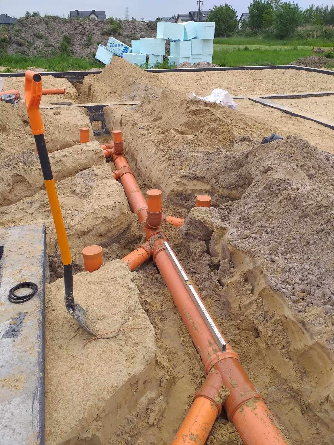 Pomarańczowe rury kanalizacyjne ułożone w wykopie, poziomica na rurze, łopata z pomarańczową rączką oparta o betonowy element fundamentu, widok na plac budowy i składowane płyty izolacyjne.
