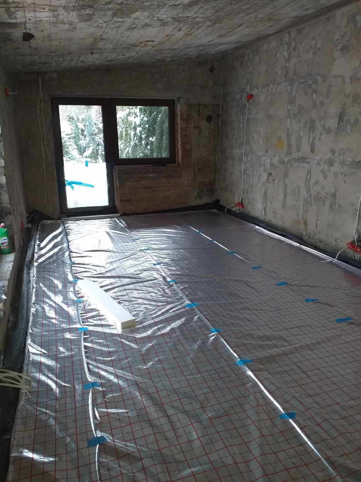Przygotowanie do instalacji ogrzewania podłogowego: mata grzewcza z siatką w czerwone kwadraty, przymocowana niebieską taśmą do podłoża, widoczne przewody elektryczne i okno z widokiem...