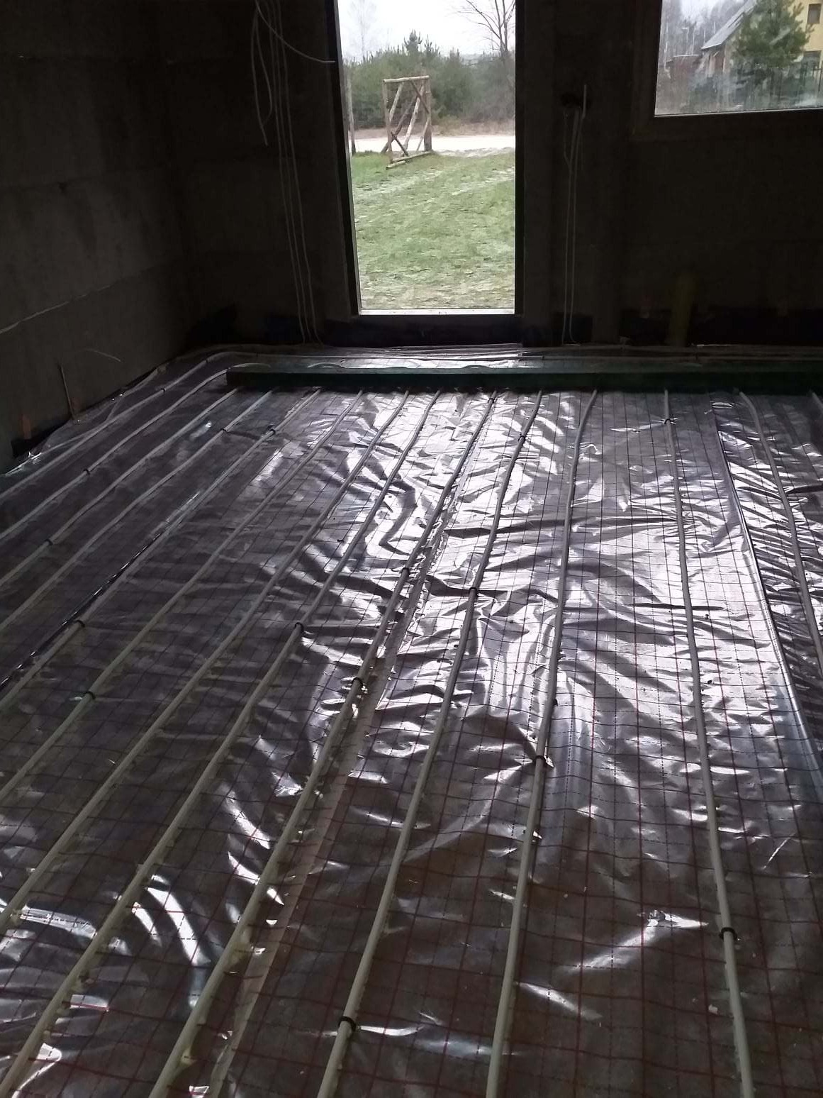 Instalacja ogrzewania podłogowego w domu w Klukach, rury rozprowadzone na folii z siatką, widok przez otwarte drzwi na zewnątrz.