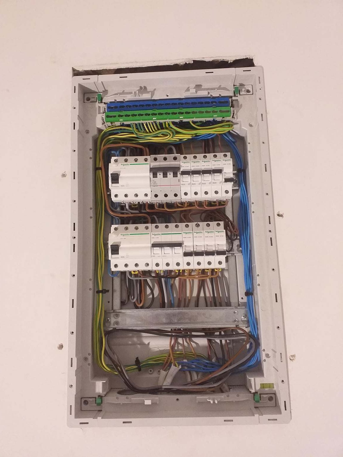 Rozdzielnica elektryczna Schneider zamontowana we wnęce ściany, widoczne zabezpieczenia i przewody w kolorach: niebieskim, brązowym, żółto-zielonym.