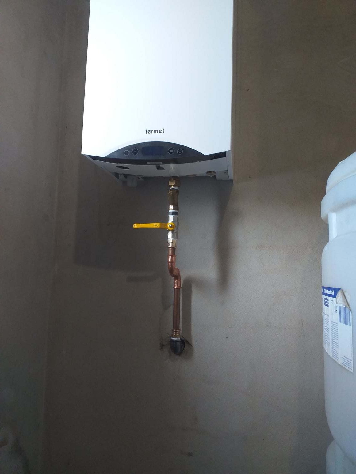 Biały wiszący kocioł gazowy marki Termet z podłączeniem miedzianymi rurami i żółtym zaworem, zamontowany na surowej ścianie.