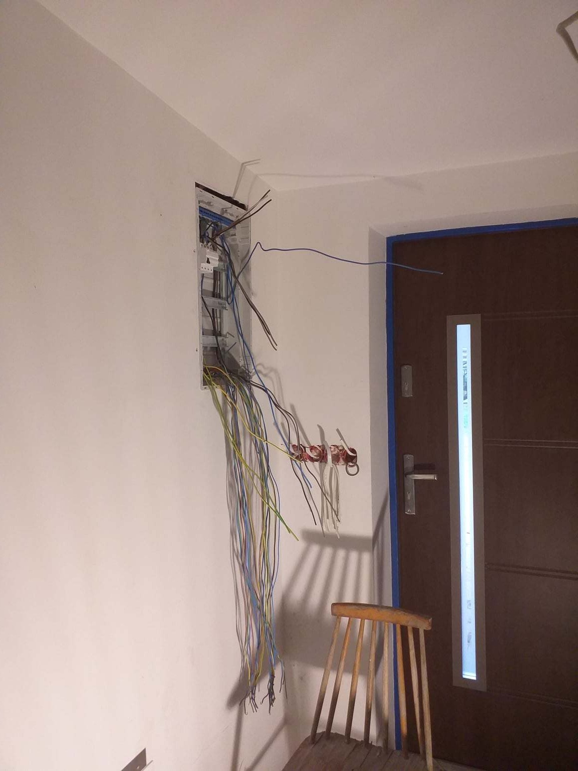 Rozdzielnia elektryczna w trakcie montażu, widoczne wystające przewody w kolorach niebieskim, żółtym i zielonym, obok drzwi wejściowe zabezpieczone taśmą malarską i drewniane krzesło.