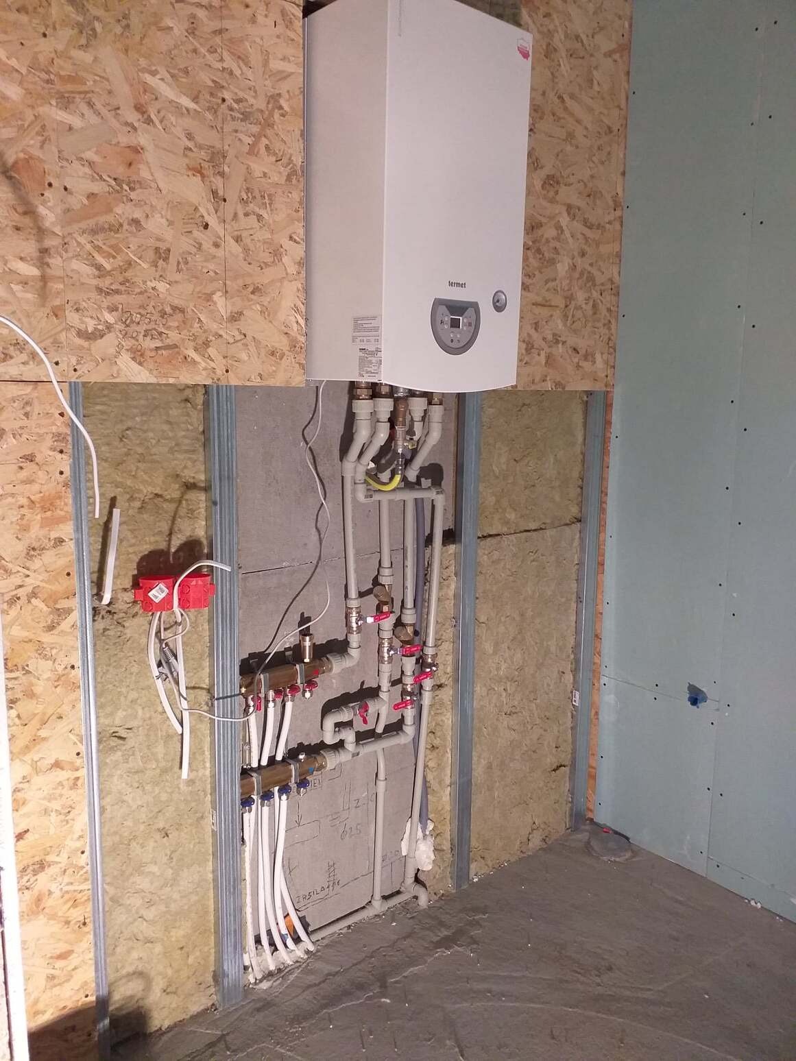 Biały wiszący piec gazowy Termet z podłączonymi rurami instalacji grzewczej i wodnej w surowym pomieszczeniu z płyt OSB i gipsowo-kartonowych, widoczne zawory i kształtki.