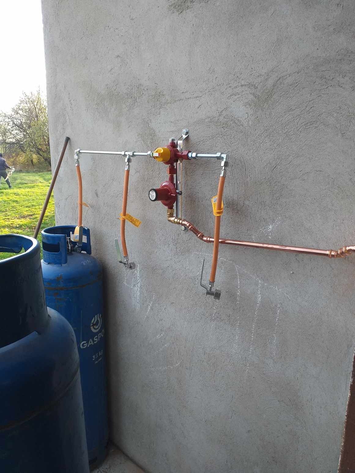 Zewnętrzna instalacja gazowa z dwoma reduktorami, rurami miedzianymi i elastycznymi przewodami, zamontowana na szarej ścianie budynku. Dwie niebieskie butle gazowe widoczne w lewym dolnym rogu.