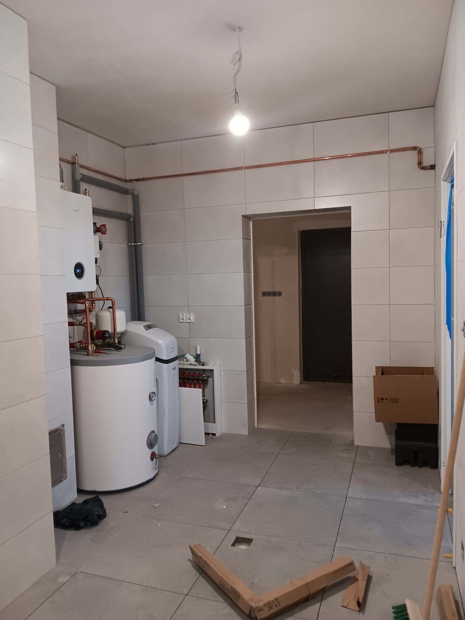 Nowoczesna kotłownia w trakcie wykańczania: biały zasobnik ciepłej wody, piec gazowy kondensacyjny, zmiękczacz wody i rozprowadzona instalacja miedziana na tle jasnych płytek.